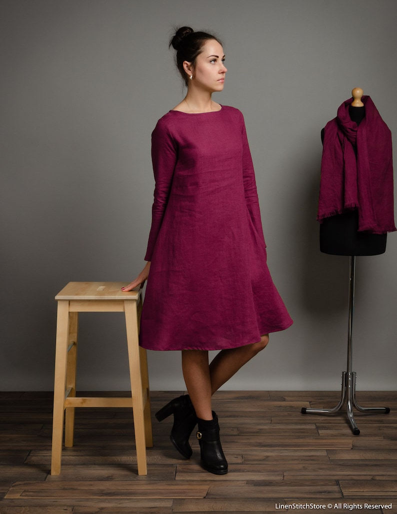 May include: A long-sleeved, knee-length magenta dress with a boat neckline and a flared skirt silhouette. The dress is made of a textured fabric and is displayed alongside a matching magenta scarf draped over a black mannequin form.