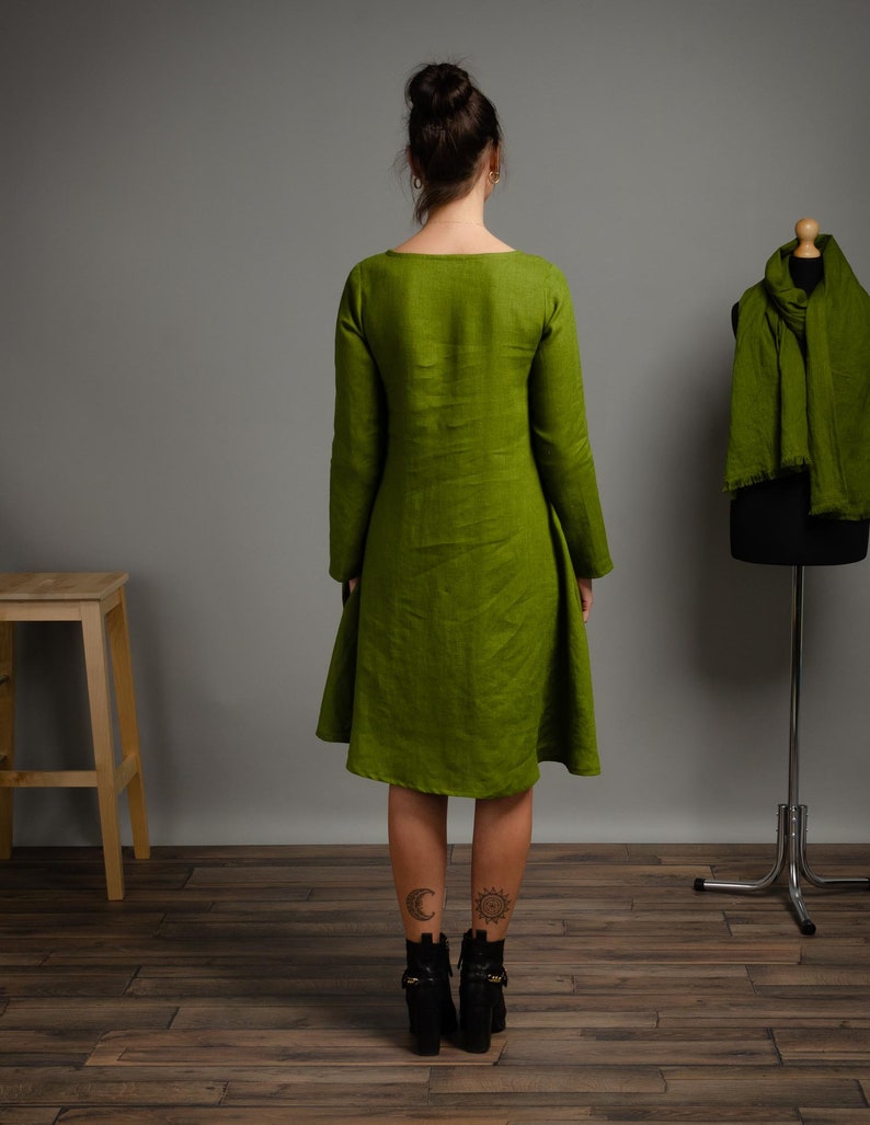 May include: A long-sleeved, green linen dress with a scoop neck and a slightly flared skirt. The dress is shown on a model with black ankle boots.