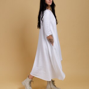 May include: A white linen dress with a loose, flowing silhouette. The dress has a round neckline and three-quarter sleeves. The model is wearing the dress with beige ankle boots.