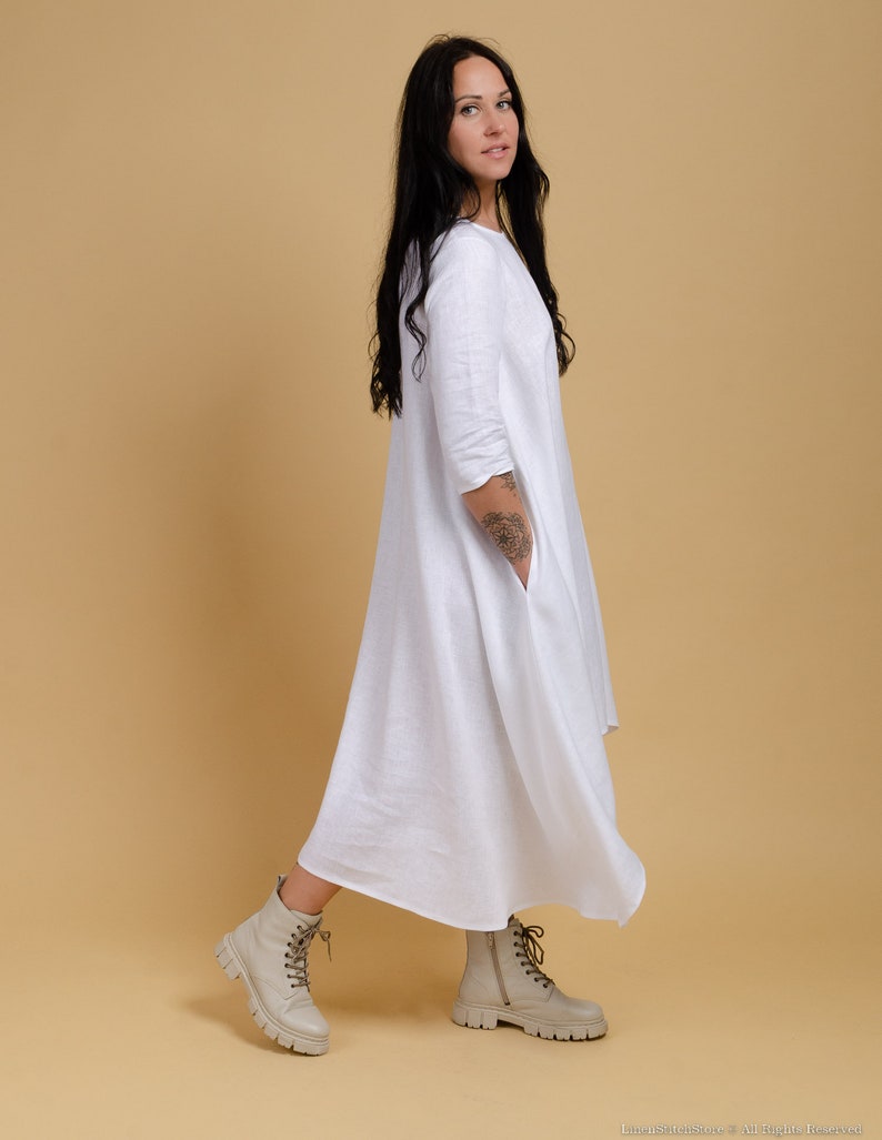 May include: A white linen dress with a loose, flowing silhouette. The dress has a round neckline and three-quarter sleeves. The model is wearing the dress with beige ankle boots.