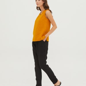May include: A woman is wearing a sleeveless, mustard yellow top and black trousers. She is standing with her hand in her pocket and looking to the side.