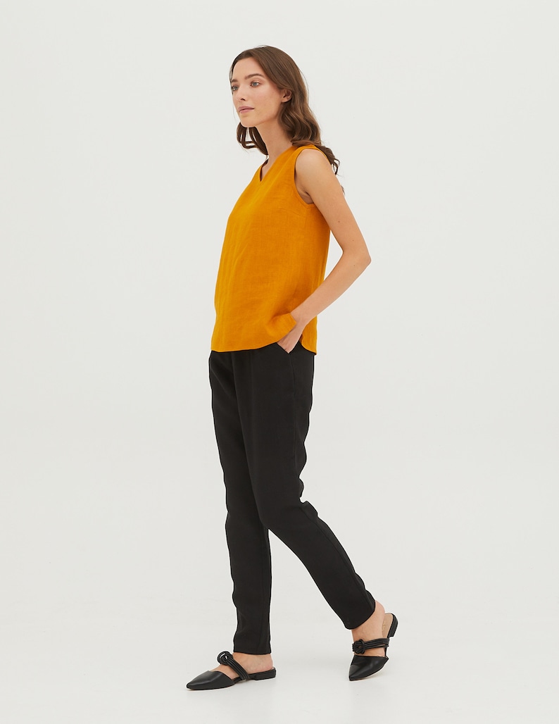 May include: A woman is wearing a sleeveless, mustard yellow top and black trousers. She is standing with her hand in her pocket and looking to the side.