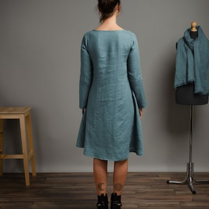 May include: A teal linen dress with long sleeves and a slightly flared skirt. The dress is shown on a model from the back, with the model wearing black ankle boots.