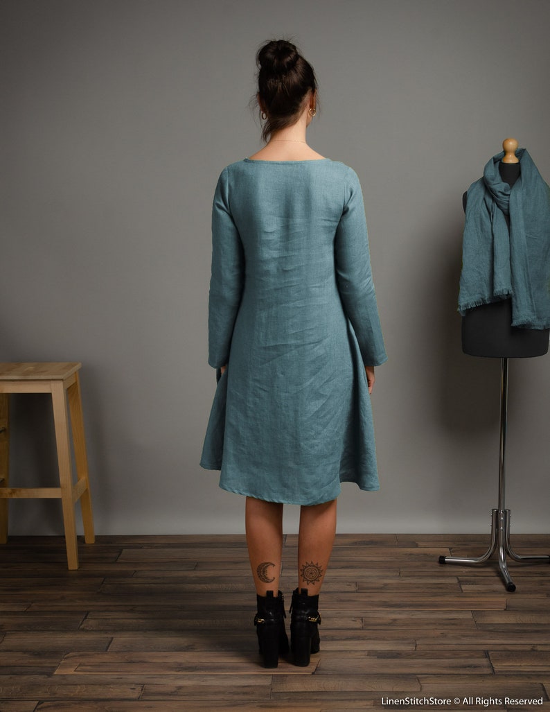 May include: A teal linen dress with long sleeves and a slightly flared skirt. The dress is shown on a model from the back, with the model wearing black ankle boots.