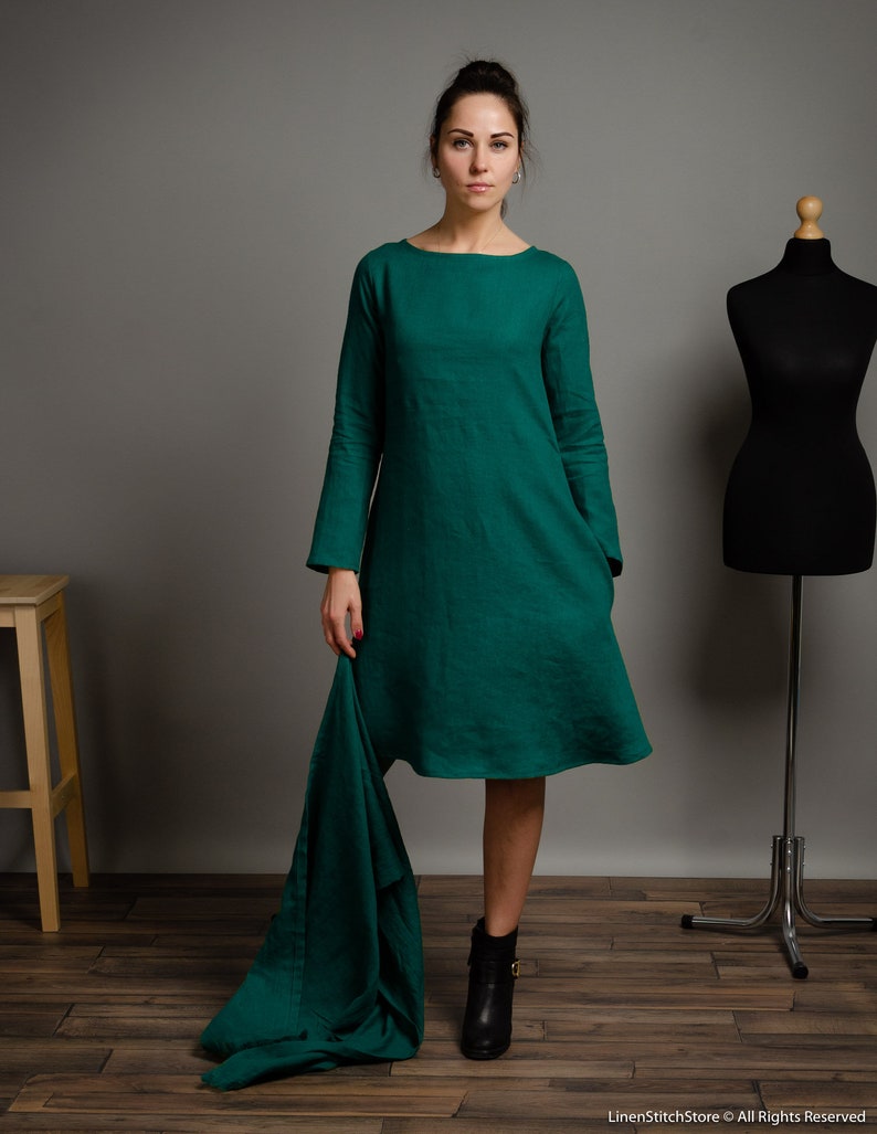 May include: A woman wearing a long-sleeved, emerald green linen dress with a boat neck. The dress has a slightly flared skirt and is paired with black ankle boots.