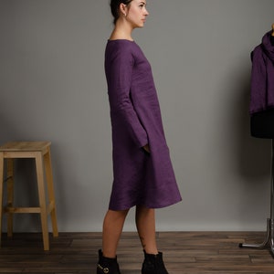 May include: A woman wearing a long-sleeved, purple linen dress with a loose fit. She is wearing black ankle boots with gold accents. The dress has a scoop neckline and a slightly flared hem.