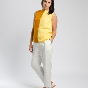 May include: A woman wearing a sleeveless top with a yellow and mustard color block design. The top has a high neckline and a relaxed fit. She is also wearing white linen pants and white sneakers.