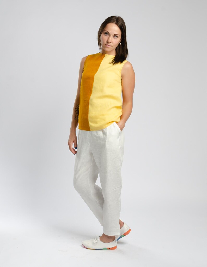 May include: A woman wearing a sleeveless top with a yellow and mustard color block design. The top has a high neckline and a relaxed fit. She is also wearing white linen pants and white sneakers.