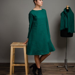 May include: A woman wearing a teal green linen dress with long sleeves and a boat neck. She is standing on a wooden floor with a wooden stool beside her.