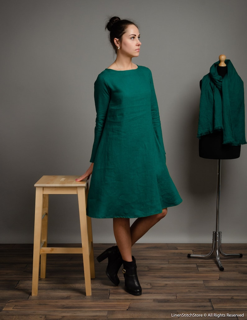 May include: A woman wearing a teal green linen dress with long sleeves and a boat neck. She is standing on a wooden floor with a wooden stool beside her.