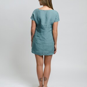 Op de afbeelding: Een teal linnen jurk met korte mouwen en een ronde hals. De jurk is aangesloten en valt net boven de knie. Het model draagt de jurk met een paar beige sneakers.