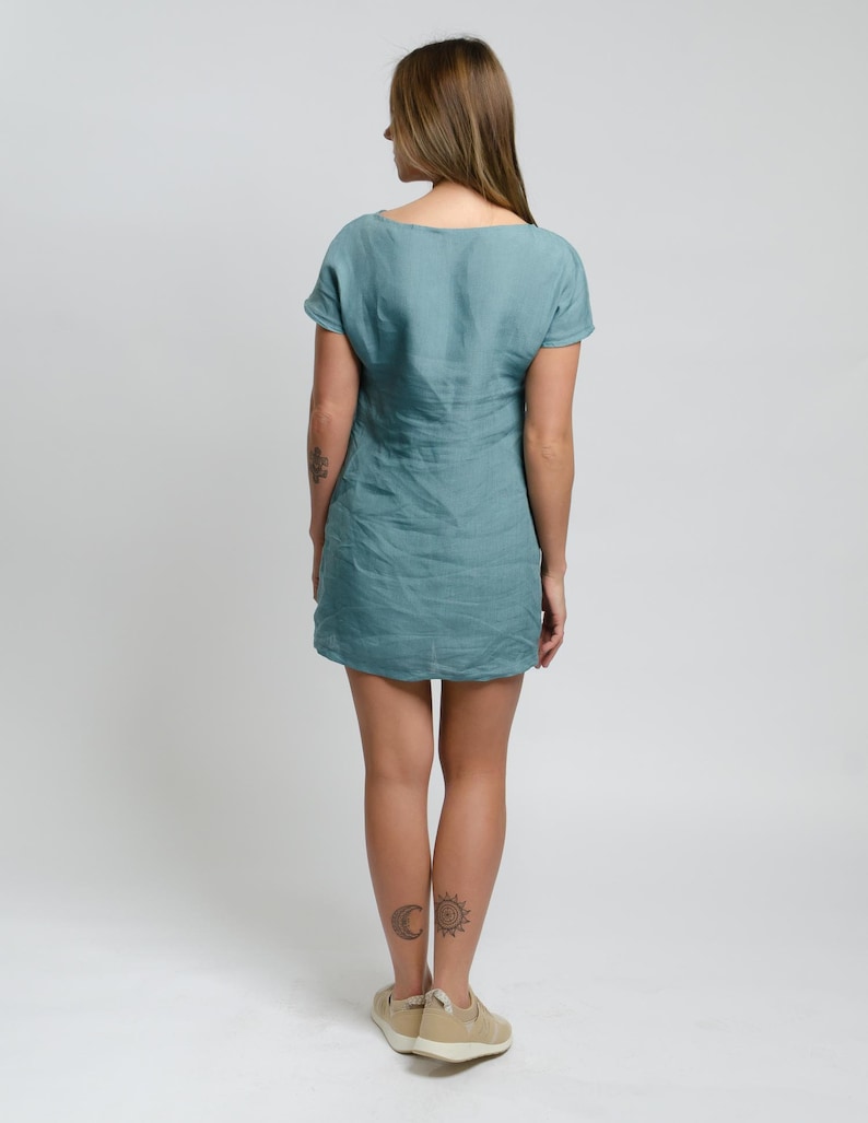 Op de afbeelding: Een teal linnen jurk met korte mouwen en een ronde hals. De jurk is aangesloten en valt net boven de knie. Het model draagt de jurk met een paar beige sneakers.