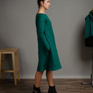 May include: A woman wearing a long, emerald green linen dress with long sleeves and a round neckline. She is wearing black ankle boots with gold accents. The dress has a loose fit and falls to her knees.