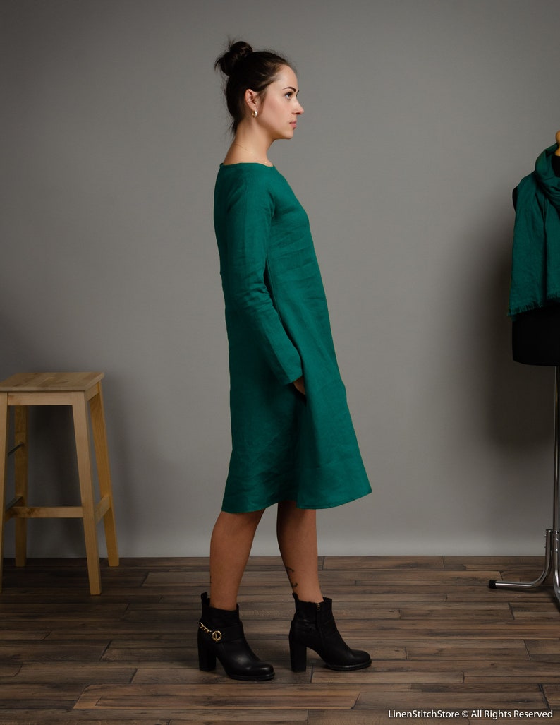 May include: A woman wearing a long, emerald green linen dress with long sleeves and a round neckline. She is wearing black ankle boots with gold accents. The dress has a loose fit and falls to her knees.