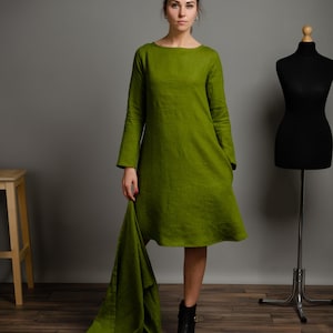 May include: A woman wearing a long-sleeved, olive green linen dress with a loose fit and a slightly flared hem. She is holding a matching green linen scarf in her hand. She is wearing black ankle boots.