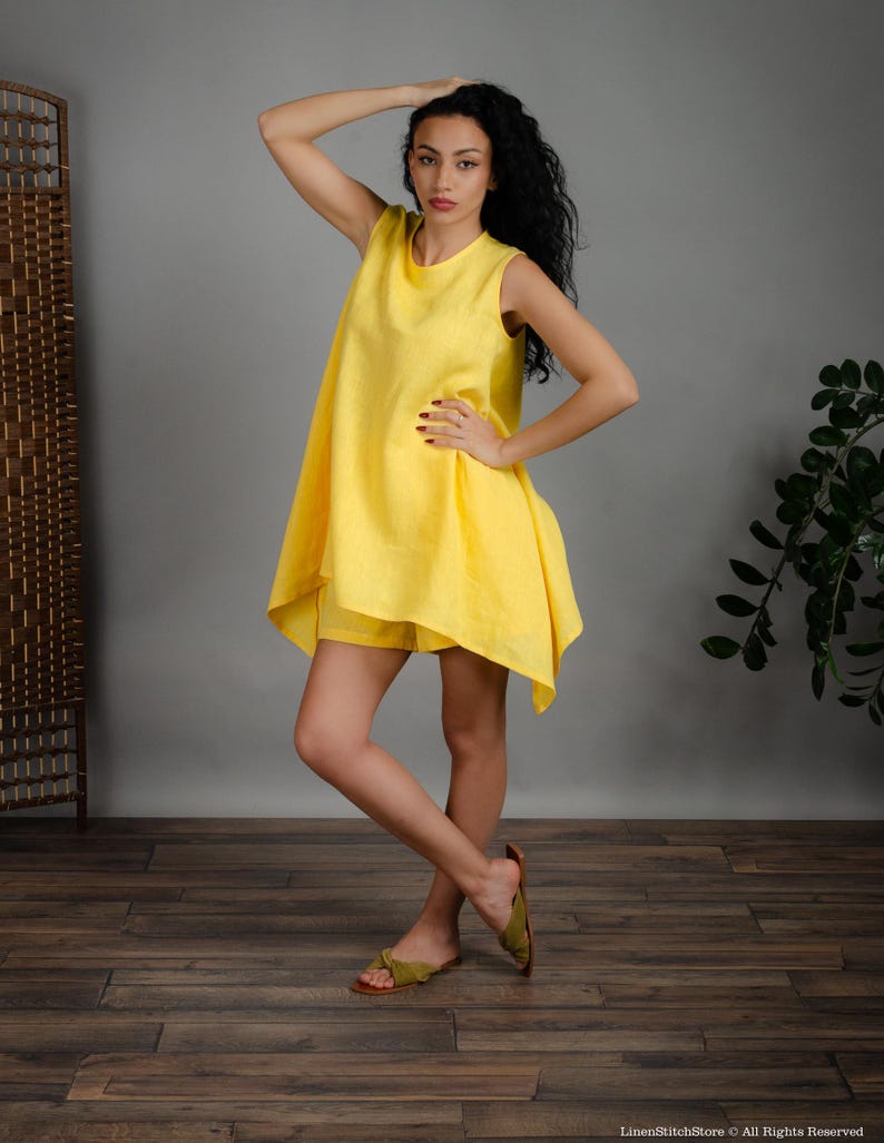 May include: A woman wearing a bright yellow linen dress with a sleeveless design and a slightly asymmetrical hemline. The dress has a relaxed fit and is paired with matching yellow linen shorts. The woman is standing on a wooden floor and is wearing green sandals.