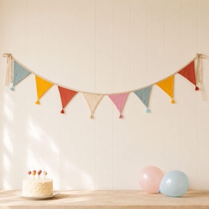 May include: A decorative bunting banner featuring nine triangular fabric flags in muted shades of blue, yellow, red, cream, and pink, each finished with a small tassel. The banner is suspended by beige fabric ties at both ends.