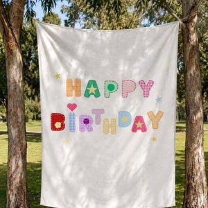 May include: A white fabric banner featuring the text "HAPPY BIRTHDAY" in large, colourful, patterned letters. Each letter displays a unique design, including polka dots, gingham, and stars. Small star and heart icons are interspersed among the text.