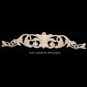 May include: A carved wooden applique with a floral design. The applique features a central motif of a stylized flower with leaves and grapes. The applique is made of light-colored wood and has a smooth finish.