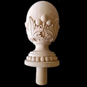 May include: A carved wooden finial with an egg-shaped top, adorned with leaf and bud carvings. The finial sits atop a pedestal base with a series of small, rounded beads. A cylindrical post extends from the base. The wood has a natural, light color.