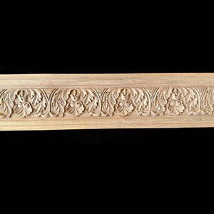 May include: A section of unfinished wood trim with a carved floral and leaf pattern. The trim is light brown and features detailed relief carvings of stylized flowers and foliage. The design is symmetrical and repeats along the length of the trim.