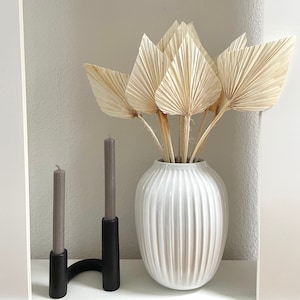 May include: A white ceramic vase with vertical ridges holds a bouquet of dried palm leaves. Two gray candles are in a black candle holder.