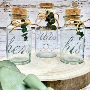 May include: Three clear glass jars with cork lids and twine tied around the necks. The jars are labeled "hers", "ours", and "his" in a silver script font. Each jar has a small sprig of eucalyptus leaves inside.