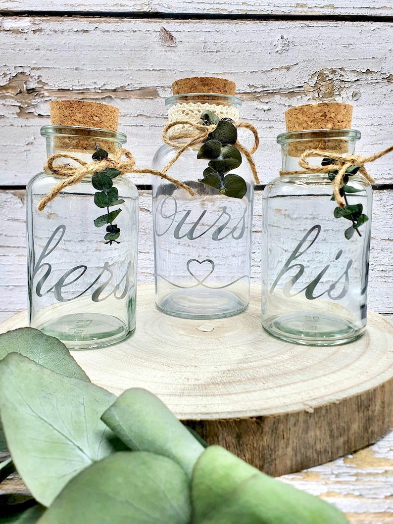 May include: Three clear glass jars with cork lids and twine tied around the necks. The jars are labeled "hers", "ours", and "his" in a silver script font. Each jar has a small sprig of eucalyptus leaves inside.