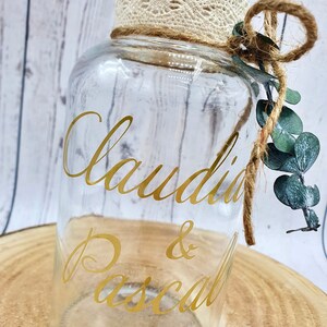 May include: A clear glass jar with a cork lid and a lace trim. The jar has a gold vinyl sticker with the names "Claudia & Pascal" written in cursive.
