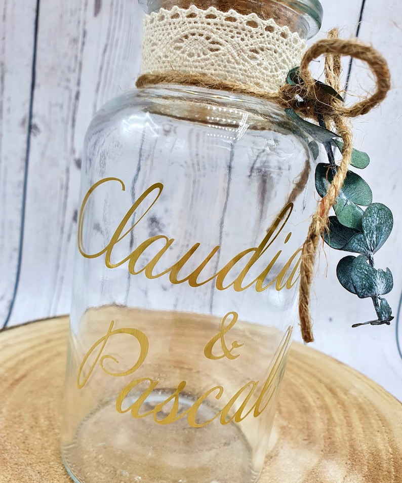 May include: A clear glass jar with a cork lid and a lace trim. The jar has a gold vinyl sticker with the names "Claudia & Pascal" written in cursive.