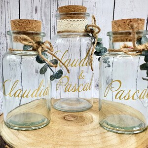 May include: Three clear glass jars with cork lids and twine accents. The jars are decorated with gold lettering that reads "Claudia & Pascal" and "Pascal".