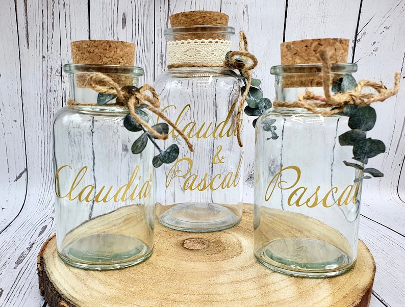 May include: Three clear glass jars with cork lids and twine accents. The jars are decorated with gold lettering that reads "Claudia & Pascal" and "Pascal".