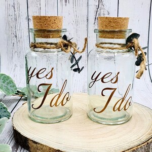May include: Two clear glass jars with cork lids and twine accents. Each jar has the words "yes I do" written in gold lettering.