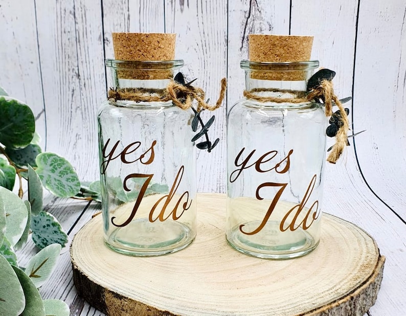 May include: Two clear glass jars with cork lids and twine accents. Each jar has the words "yes I do" written in gold lettering.