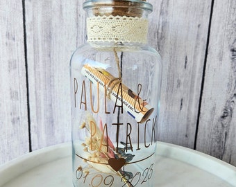 May include: A clear glass jar with a cork lid and a lace ribbon around the top. The jar contains dried flowers, a rolled-up piece of paper, and a heart-shaped charm. The jar is decorated with the names "Paula & Patrick" and the date "01.09.2023".