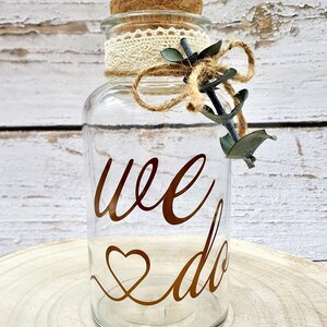 May include: A clear glass jar with a cork lid and a lace ribbon tied around the top. The jar has the words "we do" written in gold lettering with a heart symbol. The jar is decorated with a small sprig of eucalyptus.