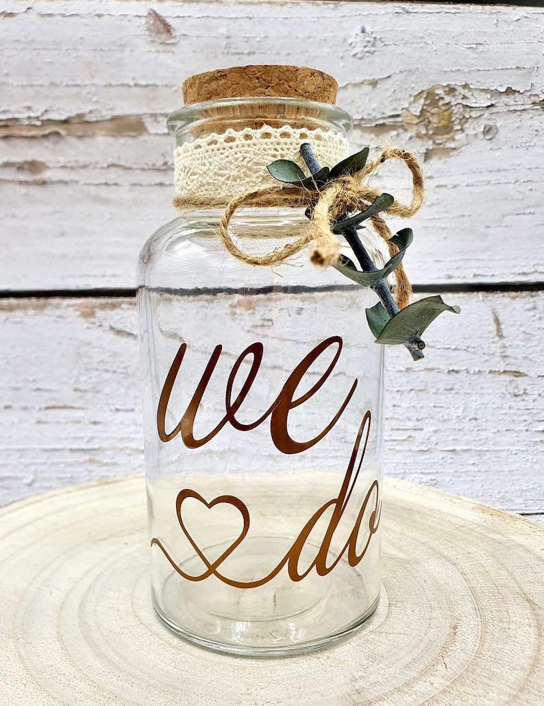 May include: A clear glass jar with a cork lid and a lace ribbon tied around the top. The jar has the words "we do" written in gold lettering with a heart symbol. The jar is decorated with a small sprig of eucalyptus.