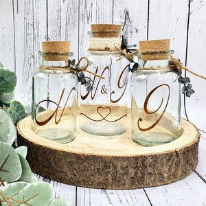 May include: Three clear glass jars with cork lids and twine accents. The jars are decorated with gold lettering, one with the letters "H & G", one with the letter "N", and one with the letter "G". The jars are sitting on a wooden slice.