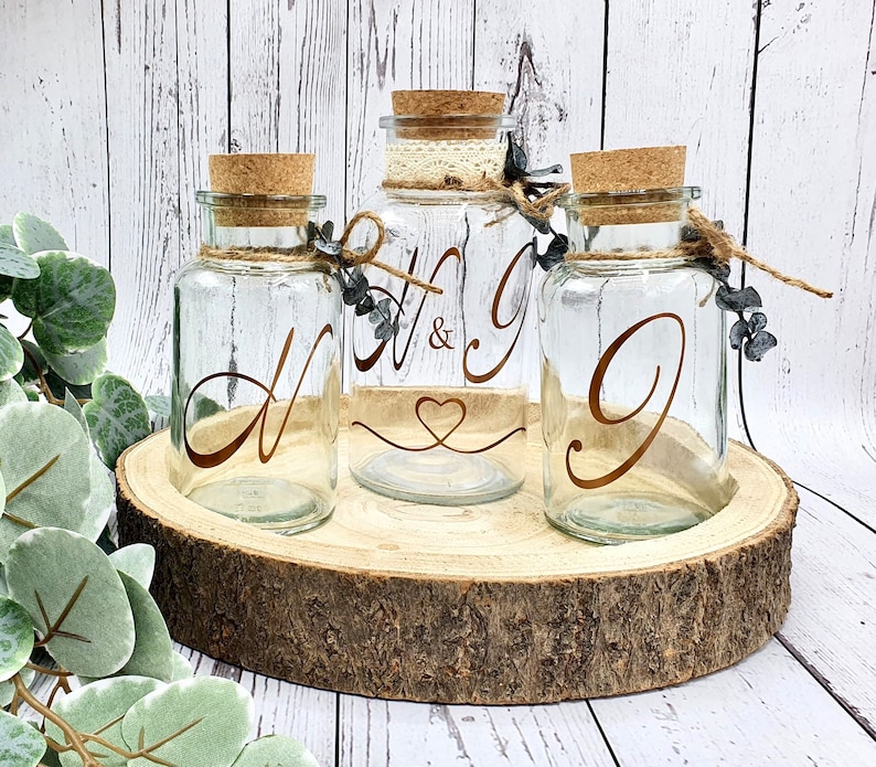 May include: Three clear glass jars with cork lids and twine accents. The jars are decorated with gold lettering, one with the letters "H & G", one with the letter "N", and one with the letter "G". The jars are sitting on a wooden slice.