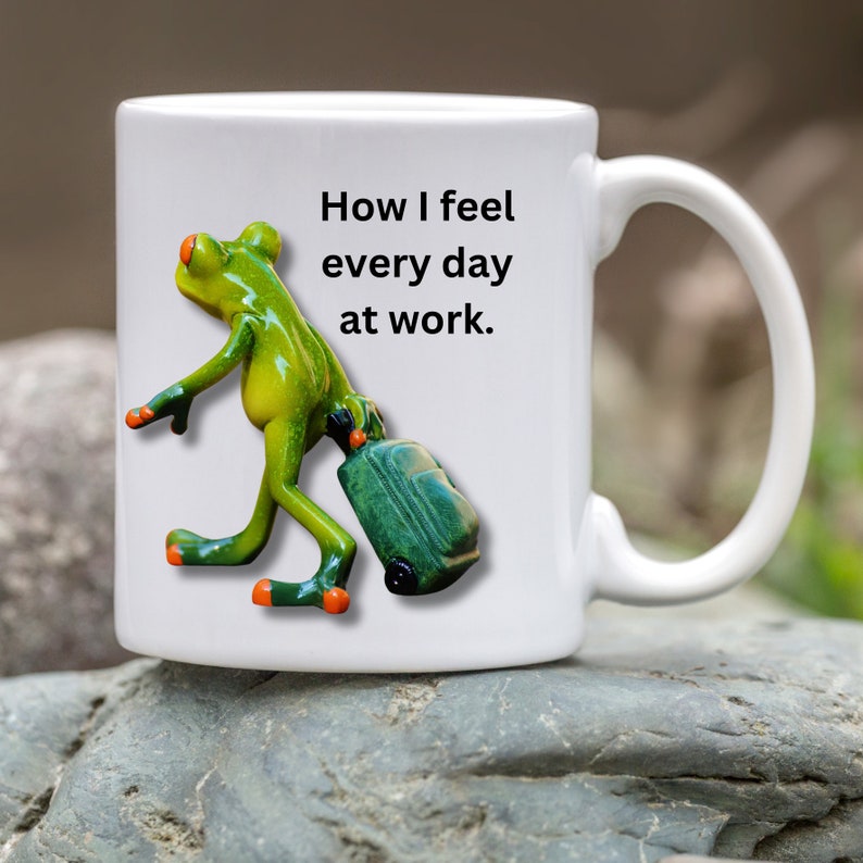 Puede incluir: Taza de cer&aacute;mica blanca con una figura de rana verde que lleva una maleta verde. La taza tiene el texto "How I feel every day at work." La rana tiene detalles en naranja en los pies y los ojos.