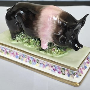 May include: A ceramic pig figurine featuring a black and pink coat, standing on a green grassy base. The base is rectangular with a white border decorated with a colorful floral pattern and gold trim along the edges of the platform.