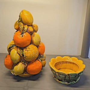 May include: A ceramic fruit topiary sculpture featuring a stack of yellow lemons and orange oranges with green leaves, accompanied by a separate matching ceramic bowl with a scalloped rim and a textured basket-weave base design.