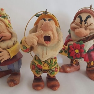 Op de afbeelding: Drie handbeschilderde dwergornamenten van hars met gedetailleerde gezichtsuitdrukkingen en kleurrijke kleding. Eén dwerg houdt een groene krans vast, een ander wijst naar de mond en de derde draagt een rode kerstster en een groene zak.