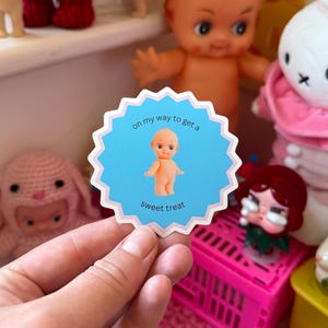 May include: A blue sticker with a scalloped edge featuring a small doll illustration and the text "on my way to get a sweet treat." The sticker is held in front of a shelf with various dolls and toys.
