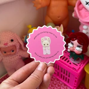 May include: A pink sticker with a scalloped edge, featuring a small white bunny in a pink blanket. The text on the sticker reads "when you are being mean to me" and "this is who you are being mean to."