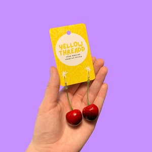 Może przedstawiać: Para kolczyk&oacute;w w kształcie wiśni na ż&oacute;łtej kartce z tekstem "Yellow Threadd Vega Maguire Creative Design".