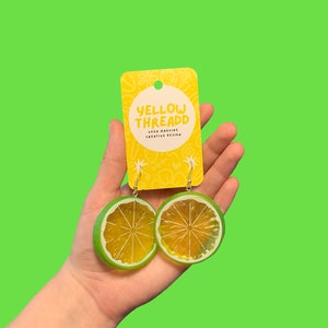 May include: A pair of lime slice earrings with green and yellow accents. The earrings are hanging from a card that says "Yellow Thread" and "Vega McGuire Creative Design".