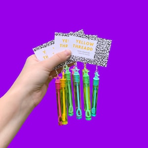 Peut inclure: Un ensemble de six baguettes à bulles colorées avec des breloques en forme d'étoile, suspendues à une carte qui indique "Yellow Threadd - Vega Maguire - Creative Design".