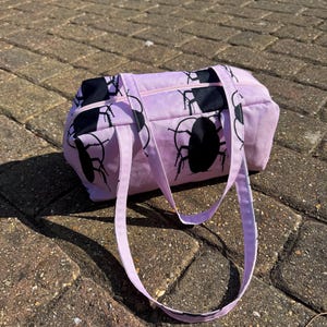 May include: A light pink rectangular handbag featuring a repeating black silhouette pattern of cockroaches. The bag has a top zipper closure and two matching light pink fabric shoulder straps attached to the top panel of the bag.
