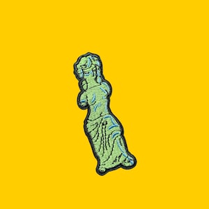 Puede incluir: Un parche bordado con la imagen de una estatua verde de una mujer con un contorno azul. La estatua est&aacute; de pie con un brazo levantado y el otro doblado en el codo.
